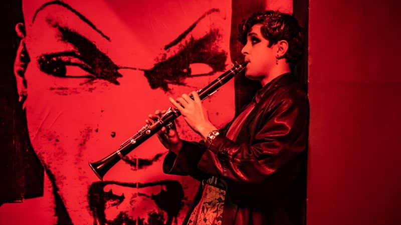 A clarinetist beside a red mural at the Kit Kat Club