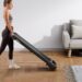 A woman carries a Merach T26B1 treadmill through a bright living room
