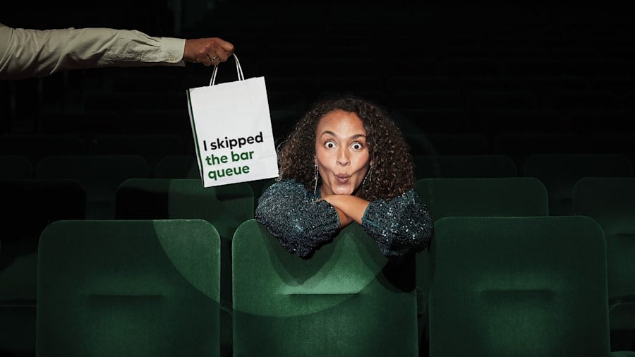 A visitor seated in a theatre auditorium with a sign about skipping the bar queue